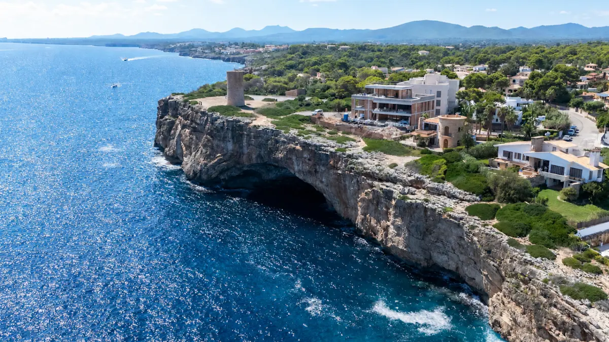 Vista aérea de un acantilado sobre una cala turquesa, con viviendas entre pinos en lo alto